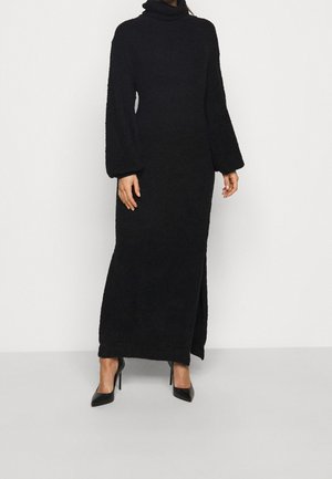 Femme portant une longue robe noire à col roulé avec des manches bouffantes et une fente sur le côté, assortie à des escarpins noirs à bouts pointus.