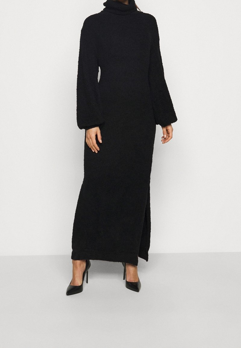 Femme portant une longue robe noire à col roulé avec des manches bouffantes et une fente sur le côté, assortie à des escarpins noirs à bouts pointus.