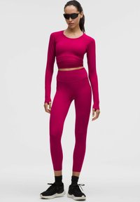 Fuksjowy krótki top z długim rękawem w zestawie z dopasowanymi legginsami. Gładki, elastyczny materiał o dopasowanym kroju. Noszony z czarnymi butami sportowymi.