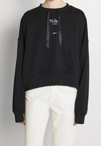 Svart kort tröja med långa ärmar, med en vit Nike-logotyp och texten "Just do it.", dragsko i huvan och slät tygstruktur.