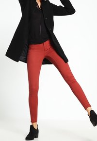 Czerwone wąskie jeansy wykonane z elastycznej tkaniny, połączone z czarnym, teksturowanym płaszczem i czarnymi botkami za kostkę, prezentujące dopasowany krój i gładką fakturę.