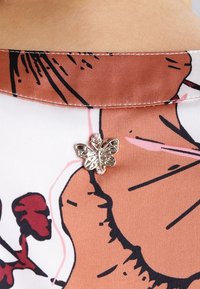 Col en fleuri de blouse en crème et marron avec une broche en argent en forme de papillon, présentant des détails complexes et une texture lisse.