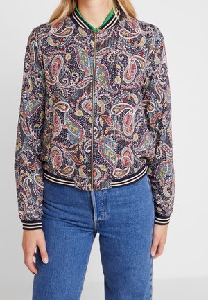 Femme portant une veste bomber multicolore à motif paisley, avec des poignets et un col rayés, associée à un jean bleu.