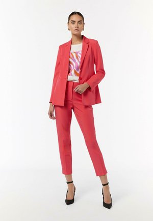 Helles pinke maßgeschneidertes Anzug mit einem strukturierten Blazer und schmalen Hosen, kombiniert mit einem cremefarbenen Oberteil mit abstrakten Mustern. Schwarze High Heels.