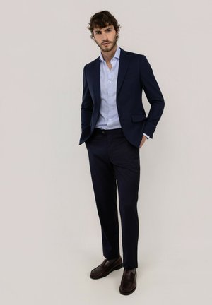 Hombre con traje azul marino, camisa azul clara, zapatos marrón oscuro, de pie con una mano en el bolsillo sobre un fondo liso.