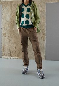 Groene lichtgewicht jas, witte trui met teal stippen, bruine broek met logo, grijze en blauwe sneakers, tegen een gestructureerde achtergrond.
