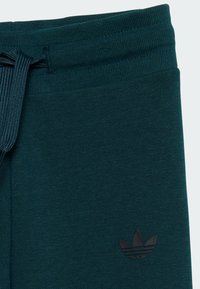Tumši zaļi kokvilnas joggeri ar ribotu jostu un aukliņu, ar mazu melnu Adidas logotipu kreisajā pusē.