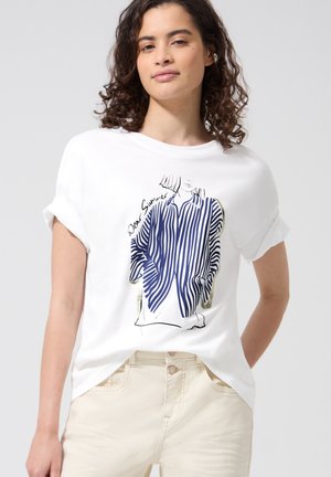 Frau mit lockigem Haar, die ein weißes T-Shirt mit einer Illustration eines blau-weiß gestreiften Hemdes und beigen Hosen trägt.