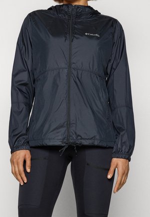 Osoba ubrana w czarną, z kapturem, wiatrówkę Columbia z zamkiem z przodu oraz czarne dopasowane spodnie na jednolitym tle.