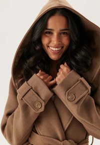 Femme souriante aux longs cheveux bruns portant un manteau à capuche marron, tenant le col près de son visage.