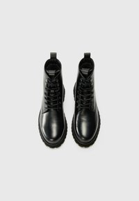Botas de cuero negras hasta el tobillo con punteras redondas, diseño de cordones y suelas de goma texturizadas. Presenta un lazo en el talón para facilitar el uso.
