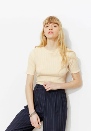 Jeune femme aux longs cheveux blonds portant un pull crème à manches courtes et un pantalon bleu marine à rayures, debout contre un mur blanc.