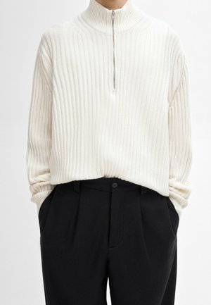 Homme portant un pull blanc côtelé à demi-fermeture éclair, rentré dans un pantalon noir taille haute, les mains dans les poches.