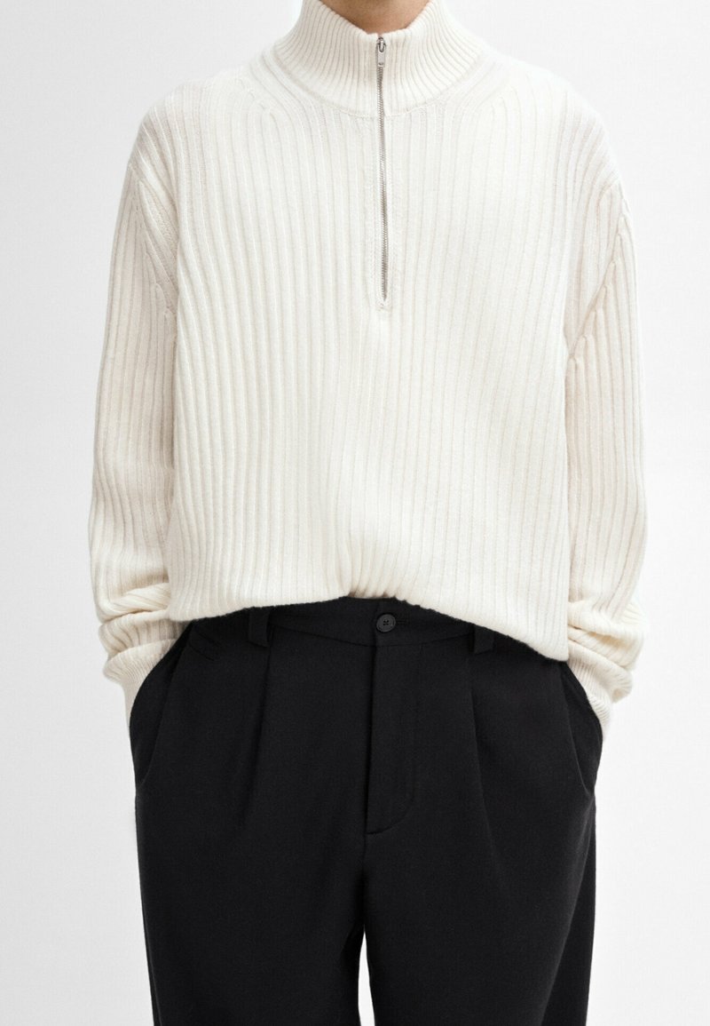 Homme portant un pull blanc côtelé à demi-fermeture éclair, rentré dans un pantalon noir taille haute, les mains dans les poches.