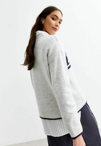 Gråstickad tröja med hög krage, marinblå detaljer och ribbad nederkant. Lager över en vit randig skjorta. Mjuk textur.