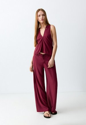 Jonge vrouw met lang haar, gekleed in een mouwloze bordeauxrode V-hals top en bijpassende broek met wijde pijpen, en zwarte slippers, staand tegen een witte achtergrond.