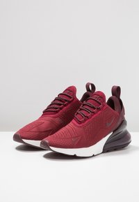 Pár červených tenisek Nike Air Max 270 s černými tkaničkami, bílými mezipodešvi a průhlednými vzduchovými polštáři na podpěrách na bílém povrchu.