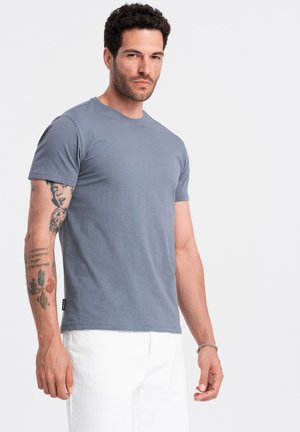 Šedé bavlněné tričko s krátkým rukávem a kulatým výstřihem, nošené s bílými kalhotami. Na ruce jsou tetování. Jednoduchý, ležérní design.