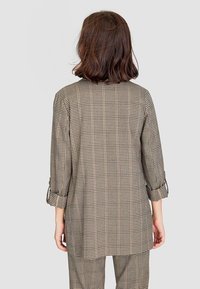 Haut à manches longues à carreaux beige et noir, avec une coupe ample, des manches retroussées et un ourlet droit vu de dos.