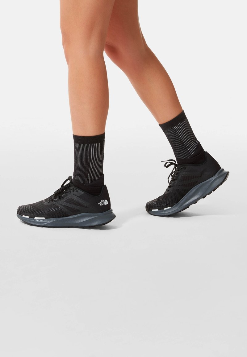 Personne portant des chaussures de course noires et des chaussettes noires rayées mi-mollet marchant sur un fond clair uni.