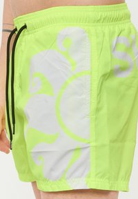 Pantaloni da bagno giallo neon con un design astratto a spirale bianca, dotati di un elastico in vita con cordino e una tasca laterale per riporre gli oggetti.