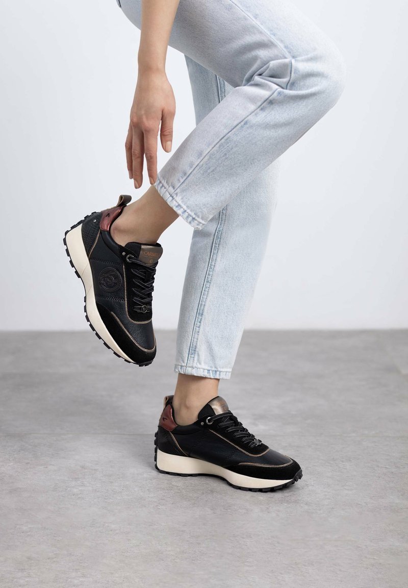 Zapatilla negra con materiales de ante y malla, que presenta una suela de color claro, un acento en el talón de color burdeos y una suela texturizada.