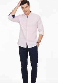 Joven con camisa de botones color rosa claro y pantalones azul marino oscuro, de pie con una mano en el bolsillo y la otra tocándose el cabello.