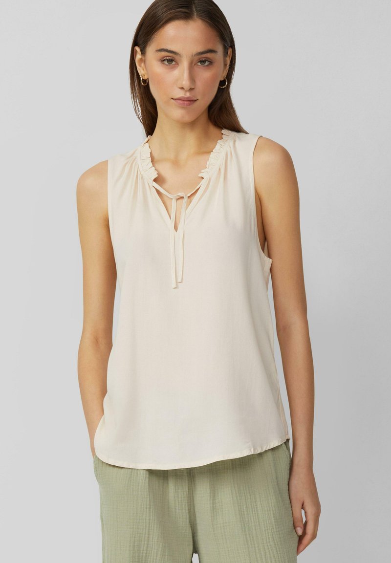 Ärmellose cremefarbene Bluse mit einem rüschierten Ausschnitt, gebundenem Detail und weichem Stoff; kombiniert mit hellgrünen strukturierten Hosen.