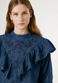 Blusa in denim con maniche arricciate e una spalla ricamata a motivo floreale in blu scuro. Presenta un collo rotondo e una vestibilità casual.