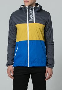 Giacca color block con un motivo a scacchi nero nella parte superiore, con sezioni gialle e blu sotto. Include un cappuccio e tasche con zip.