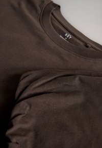Brun crew-neck langærmet skjorte lavet af blødt stof, foldet, så kraven og en del af ærmet med mærkeetiketten synlig indeni kraven vises.