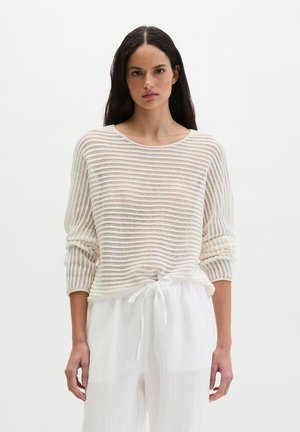 Donna con capelli lunghi e scuri che indossa un maglione bianco a maglia con maniche arrotolate e pantaloni bianchi larghi con coulisse, davanti a uno sfondo semplice.