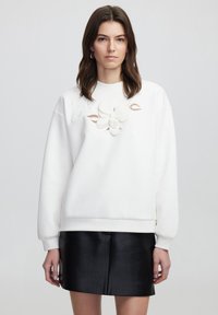 Sudadera blanca con un patrón floral bordado en el centro. Presenta mangas largas y un corte holgado, combinada con una falda de cuero negra.