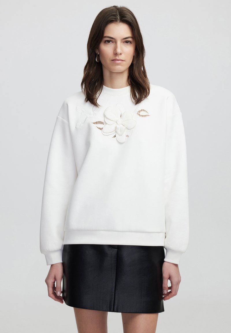 Sudadera blanca con un patrón floral bordado en el centro. Presenta mangas largas y un corte holgado, combinada con una falda de cuero negra.