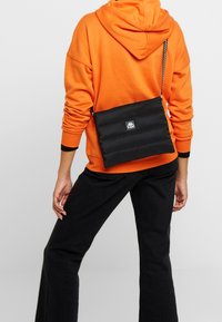 Černá prošívaná crossbody taška s logem, nastavitelným řetízkem, obdélníkovým tvarem a hladkým povrchem. Nositelná na zádech.