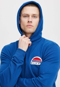 Il sudario blu con cappuccio presenta un logo ricamato rotondo con strisce rosse e blu. Realizzato in tessuto morbido, ha una vestibilità comoda e un cordino.