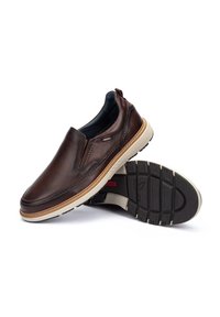 Zapatos de cuero marrón sin cordones con una superficie texturizada, suela beige en contraste y diseño de perfil bajo. Presentan una pestaña trasera para facilitar el calzado.