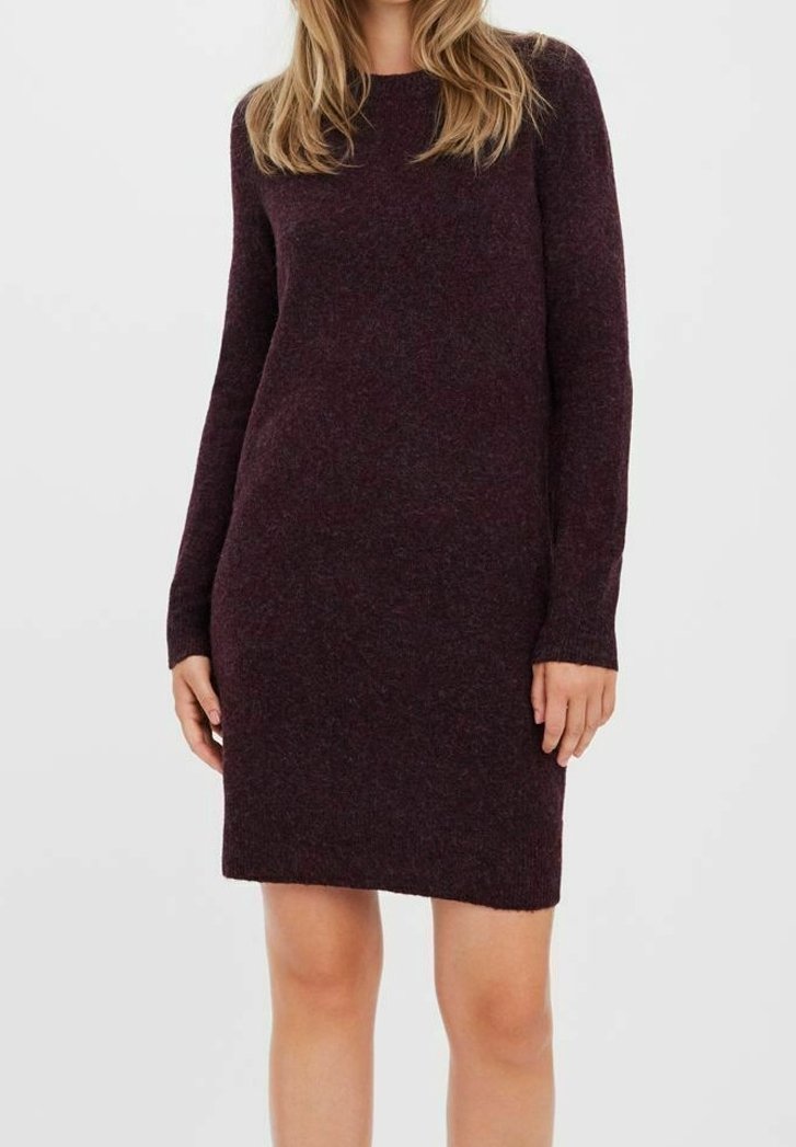 Femme portant une robe pull en tricot violet foncé à manches longues, longueur genoux, debout devant un fond uni gris clair.