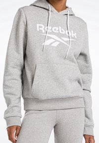 Felpa grigia con cappuccio regolabile, vestibilità oversized, tasca a marsupio e grande logo Reebok bianco sul davanti. Tessuto morbido e strutturato.