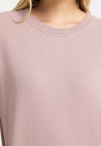 Sudadera rosa claro con cuello redondo acanalado, textura suave y corte holgado. El material parece suave y casual, adecuado para un uso relajado.