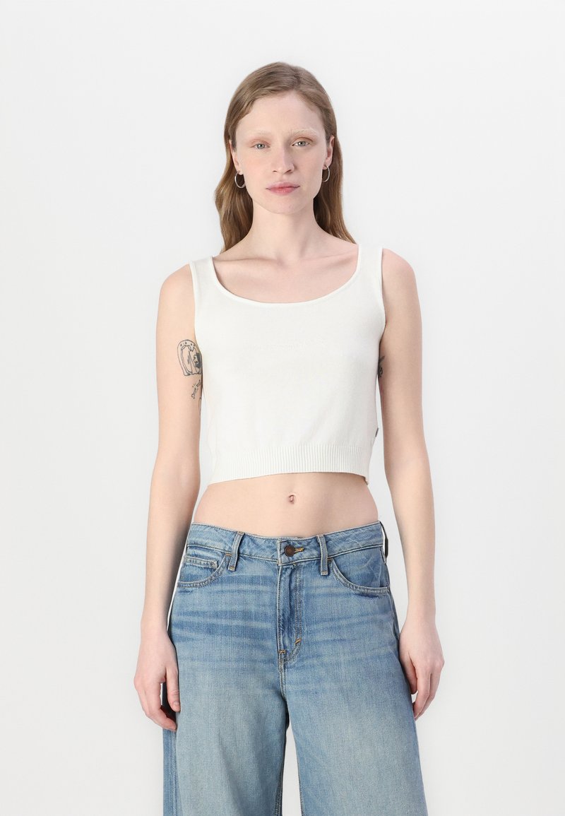 Hvit, kort tank topp med rund hals og ribbestrikket kant, parret med lyseblå denimbukser med avslappet passform og knappelukking.