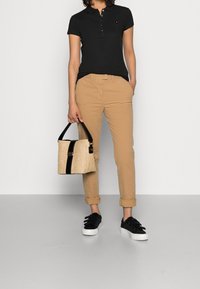 Persona che indossa una maglietta nera a maniche corte e pantaloni beige arrotolati, mentre tiene una borsa a tracolla tessuta beige e nera e scarpe nere con suole bianche.