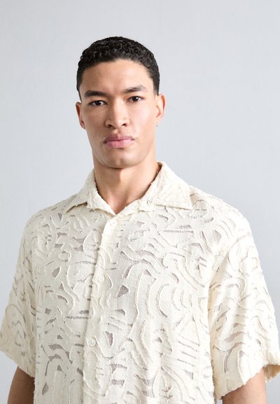 Lys beige kortermet skjorte med et teksturert, abstrakt mønster. Har spiss krage og knappelukking, laget av et mykt, pustende stoff.