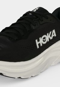 Μαύρο παπούτσι τρεξίματος Hoka με λευκή σόλα και λευκό λογότυπο Hoka στο πλάι, εμφανίζοντας λεπτομέρειες από μπροστά και από το πλάι, συμπεριλαμβανομένων των κορδονιών.