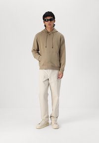 Beige hooded sweatshirt with drawstrings and front pocket, paired with light beige cargo pants and cream sneakers, model wears black sunglasses.