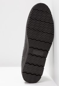 Semelle de chaussure noire avec un motif de bande de roulement en zigzag profond, positionnée verticalement contre une surface et un mur blancs.