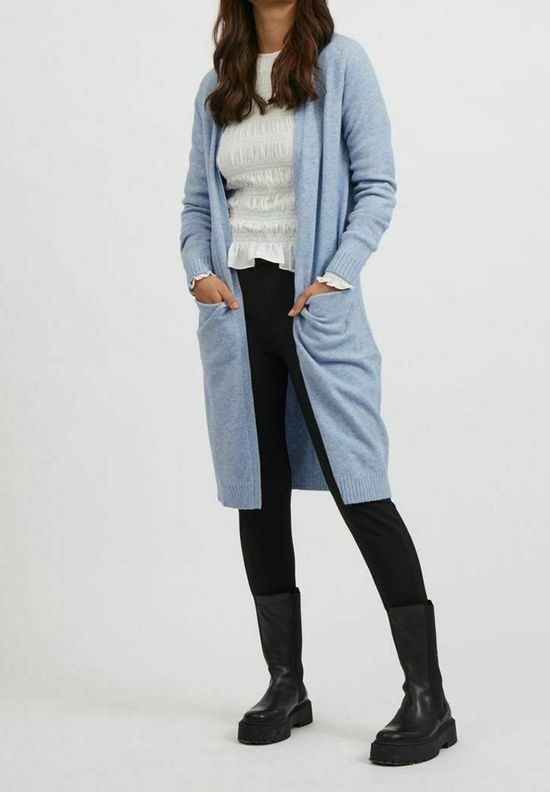 Femme portant un long cardigan bleu clair, un chemisier blanc texturé, un pantalon noir et des bottes noires épaisses, debout les mains dans les poches.