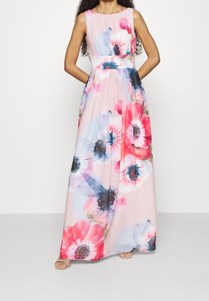 Kvinde iført ærmeløs, gulvlængde kjole med stort pink og blåt blomsterprint, stående med hænderne bag ryggen.