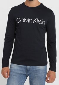 Schwarzes Langarm-T-Shirt aus Baumwolle mit einem weißen "Calvin Klein"-Logo auf der Brust. Figurbetonte Passform und runder Halsausschnitt.