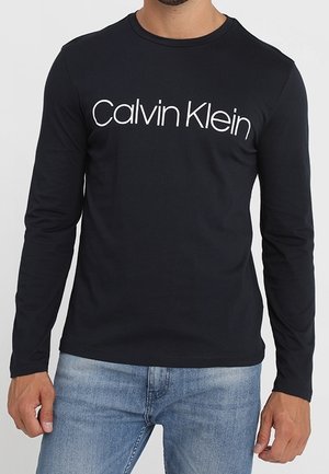 T-shirt noir à manches longues en coton, arborant un logo blanc "Calvin Klein" sur la poitrine. Coupe slim et col rond.